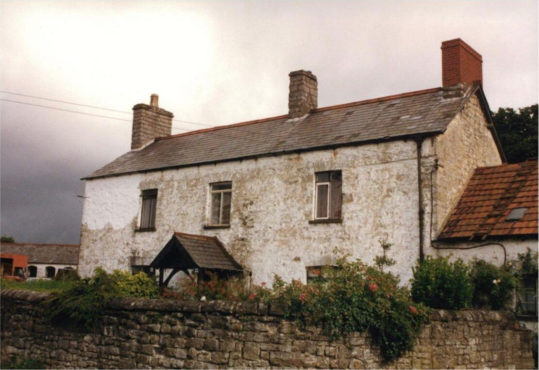 Tŷ Mawr: a two-storey limewashed stone farmhouse with three chimneys behind a rubble-stone wall, under a dark sky